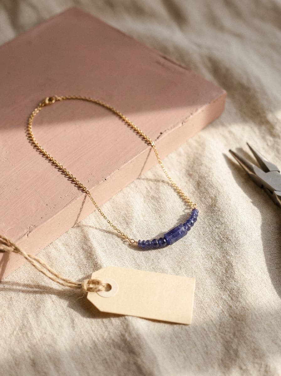 The Tanzanite Focal Necklace on its gold-filled chain, laid on natural linen beside a blank paper specimen tag — the last step before it is named and numbered.
